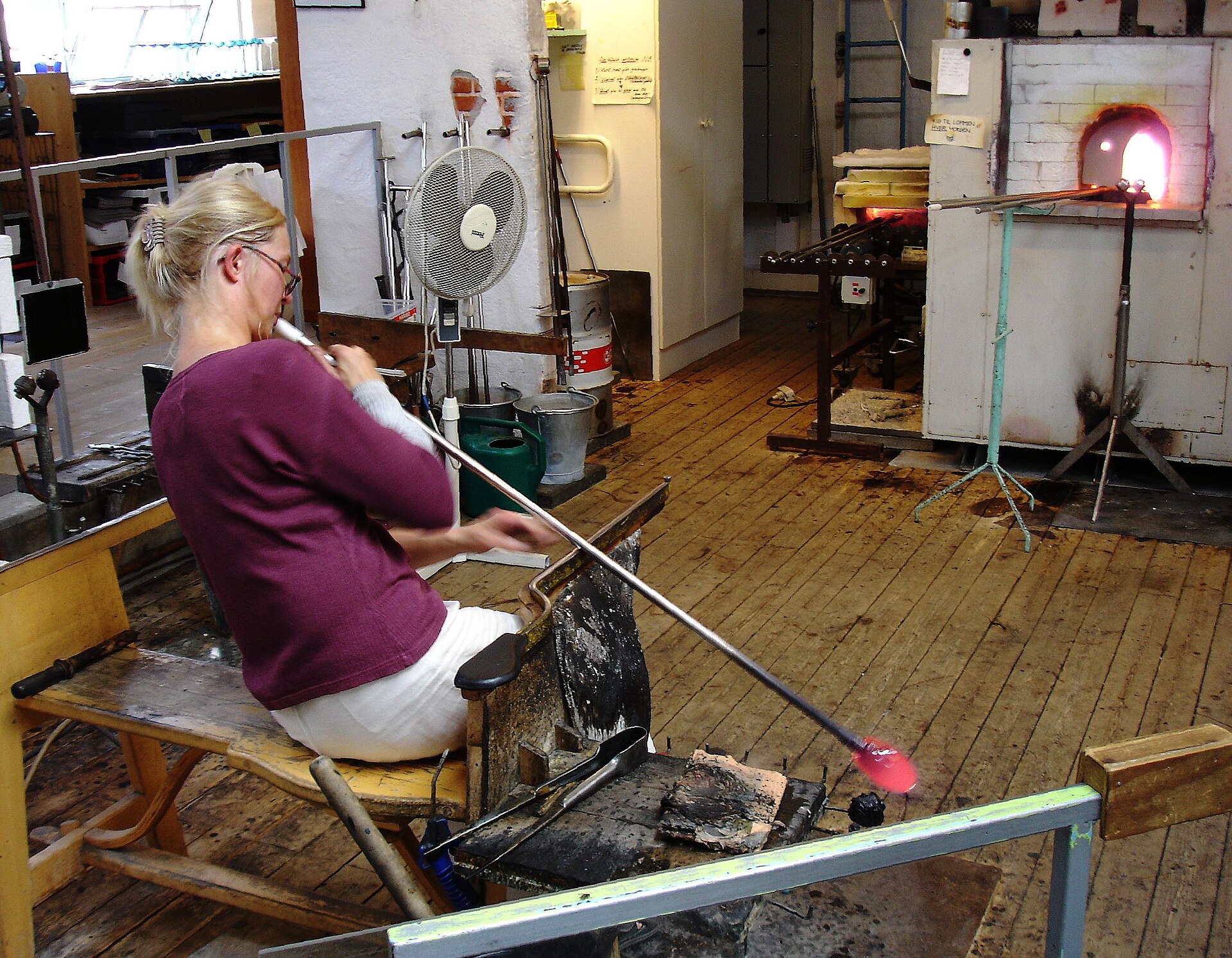 A glassworker blowing through a pipe while shaping glass at the bench